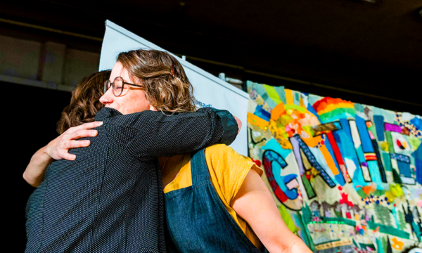 Two people embrace in front of a colourful tapestry with the word Gathering written on it