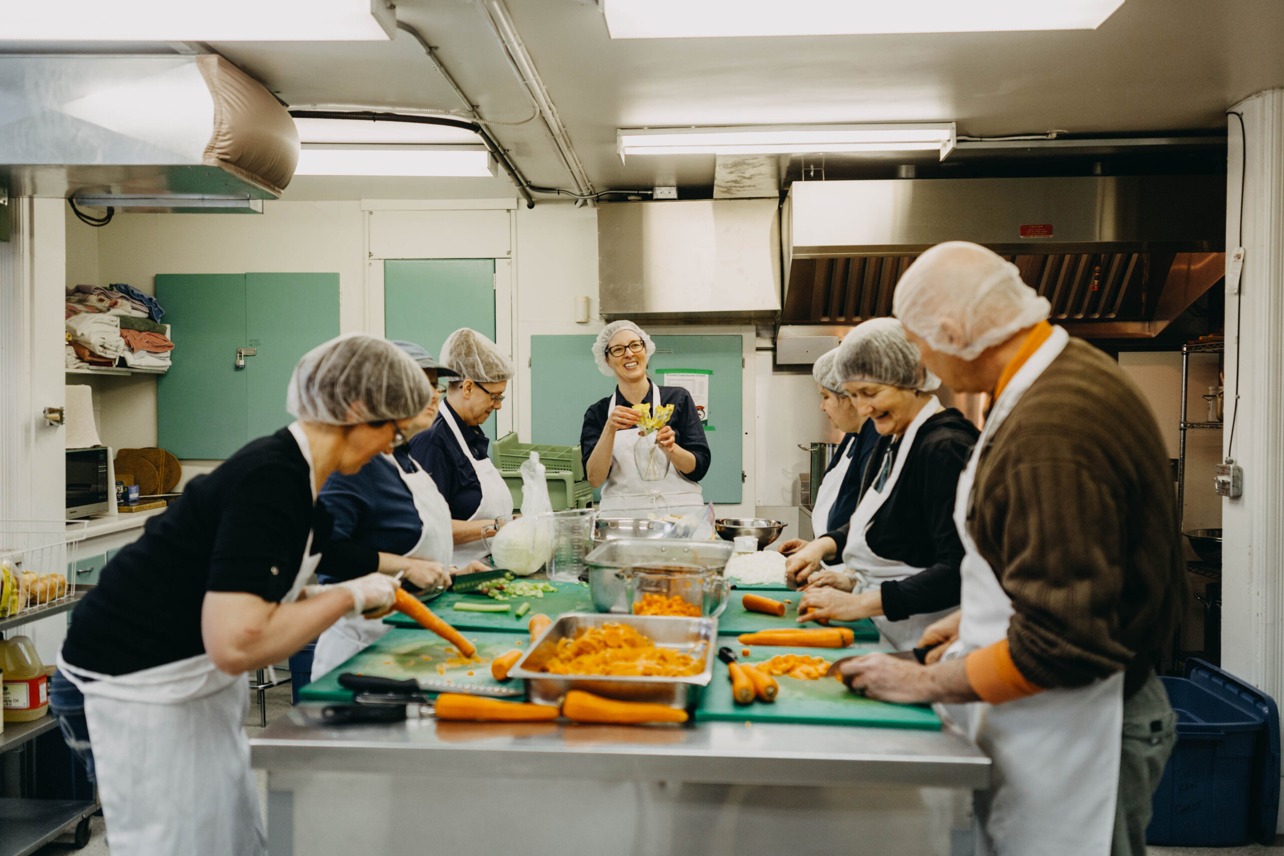 A group of people in hairnets and aprons peel and chop carrots in an industrial kitchen. They are smiling and laughing.
