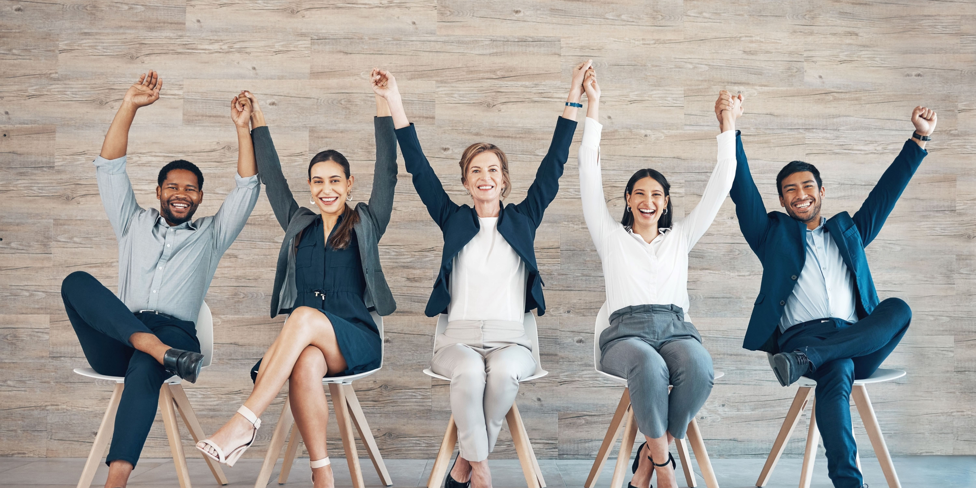 Five sitting business people sitting on chairs, holding hands with their arms raised.